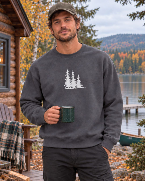 A man wearing a dark gray crewneck sweatshirt - the Spruce Trees Crewneck Sweater from Spruce and Heron - with a white pine tree graphic, standing on a lakeside deck next to a log cabin and holding a green mug.