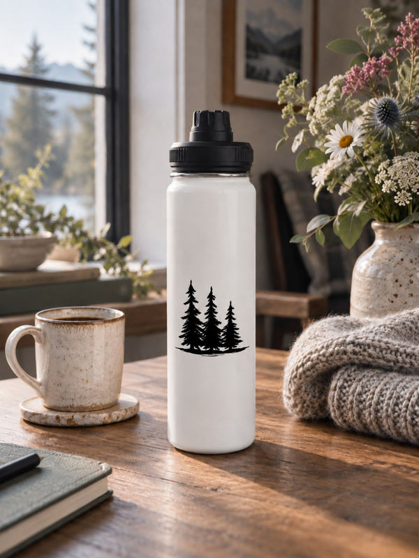 An off-white beige insulated water bottle with a black lid and a black pine tree graphic, standing on a wooden table in a cozy indoor setting with a mug, flowers, and a knitted blanket nearby - Spruce Trees Stainles Steel Water Bottle by Spruce and Heron.