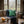 A cozy living room scene with a beige sofa, a green Fern Cushion Cover from Spruce and Heron, a navy plaid blanket, candles, a mug, and a rustic wood coffee table in front of a large bay window looking out onto a misty forest.