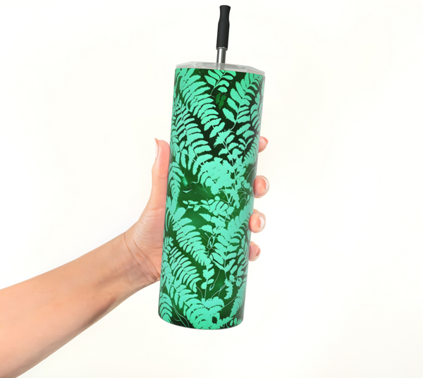 A hand holding a tall insulated tumbler with a green fern leaf pattern and a black straw, against a plain white background - Fern Tumbler - Stainless Steel - Spruce and Heron.