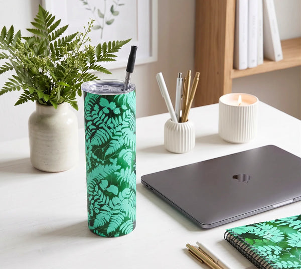 A tall insulated tumbler with a green leaf pattern and a black straw, standing on a white desk next to a closed gray laptop, pens, a candle, and a notebook - Fern Tumbler from Spruce and Heron.