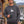 A man wearing a dark gray crewneck sweatshirt - the Spruce Trees Crewneck Sweater from Spruce and Heron -  with a white pine tree graphic, standing on a lakeside deck next to a log cabin and holding a green mug.