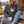 A woman sits on a wooden porch wearing a dark charcoal gray crewneck sweatshirt - nature Spruce Trees crewneck sweater from Spruce and Heron  -  with a white pine tree graphic on the chest, paired with light blue jeans, a beige knit beanie, and holding a white mug.