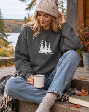 A woman sits on a wooden porch wearing a dark charcoal gray crewneck sweatshirt - nature Spruce Trees crewneck sweater from Spruce and Heron  -  with a white pine tree graphic on the chest, paired with light blue jeans, a beige knit beanie, and holding a white mug.