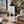 An off-white beige insulated water bottle with a black lid and a black pine tree graphic, standing on a wooden table in a cozy indoor setting with a mug, flowers, and a knitted blanket nearby - Spruce Trees Stainles Steel Water Bottle by Spruce and Heron.