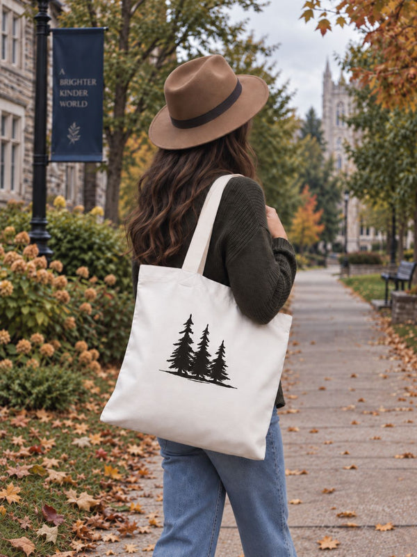 A woman wearing a brown wide-brim hat, dark green sweater, and blue jeans walks on a campus path carrying a white canvas tote bag with a black tree graphic - Spruce Trees Tote Bag from Spruce and Heron.