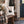 A cream canvas tote bag with black pine tree graphics sits on a rustic wooden bench in a styled entryway. The scene includes a plaid throw blanket, boots, a blue front door, and framed wall art.