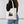 A woman from the back wearing a black long-sleeve top and blue jeans, carrying a natural canvas tote bag with a black pine tree graphic on a white paneled wall background - tote bag from Spruce and Heron.